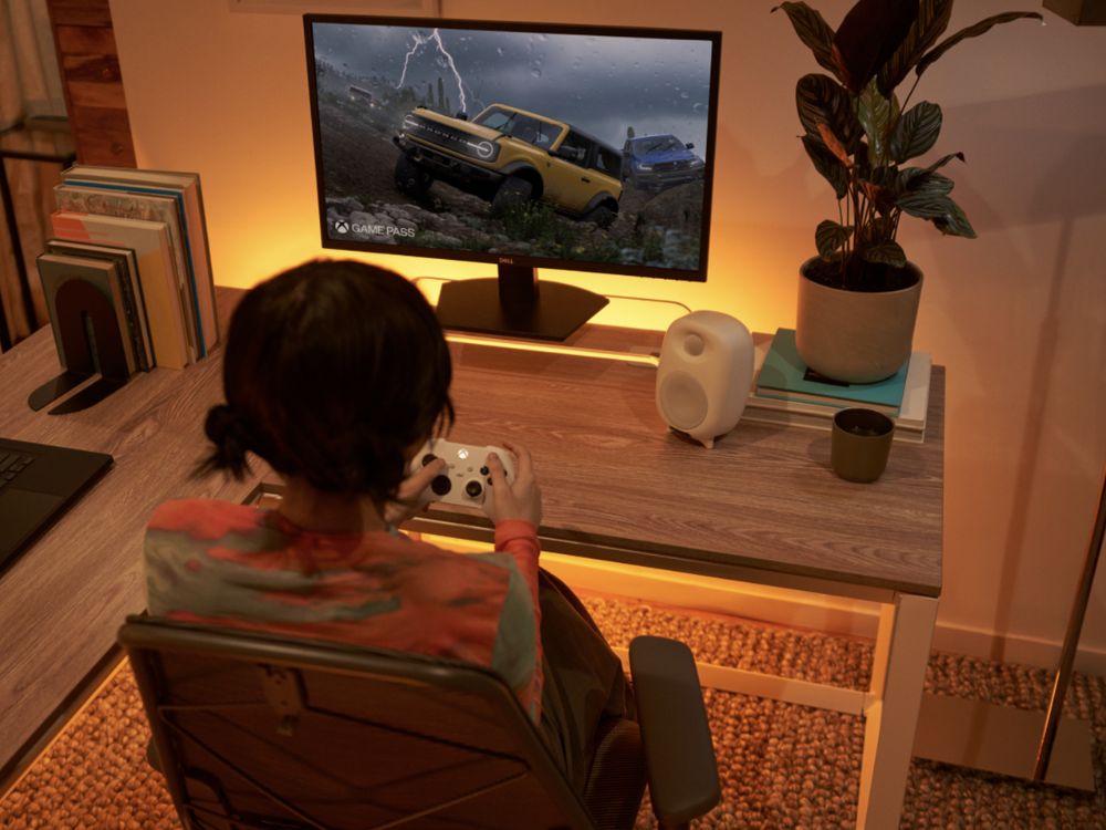 Nahaufnahme einer Person mit einem schwarzen Xbox-Controller in den Händen. Die Person spielt ein Rennspiel, das auf einem Bildschirm zu sehen ist. Auf dem Bildschirm ist ein verschwommenes Bild eines Rennwagens auf einer Strecke erkennbar. Die Hände tragen einen weißen Pullover und halten den Controller in Spielposition.